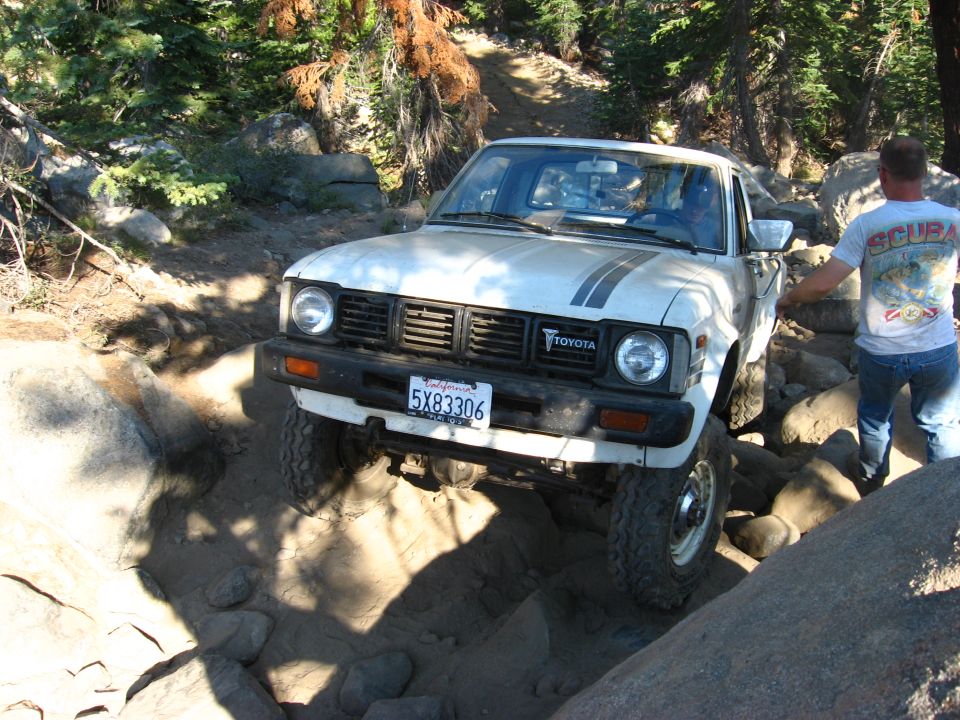 White Toyota pickup truck navigating a rocky off-road trail with a person standing nearby