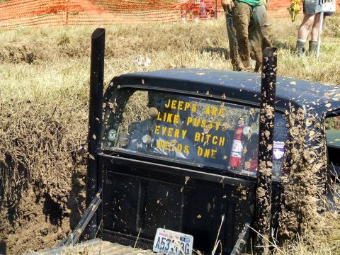 1977 Ford F-150 - A black pickup truck is stuck in the mud, with a humorous sticker on its rear window.