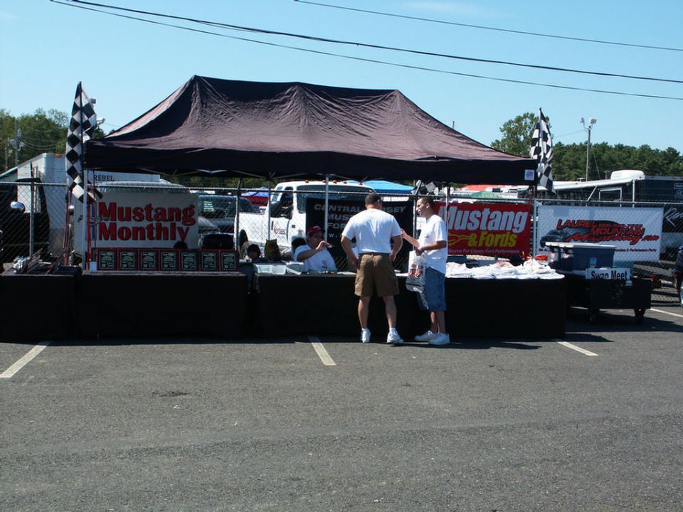 Car show tent with 'Mustang Monthly' and 'Mustang & Fords' banners, people standing around displays
