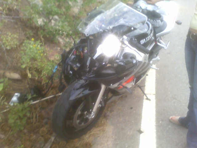 Black Suzuki sportbike with damaged front end, off the road next to some bushes. A person stands nearby
