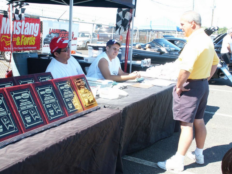 Table with car show awards, two people behind it, and a man in a yellow shirt standing nearby