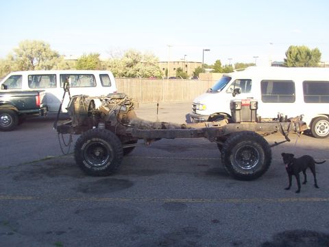 1978 Ford Bronco - The chassis of a vehicle, including the engine and suspension, is shown outdoors with vans in the background.