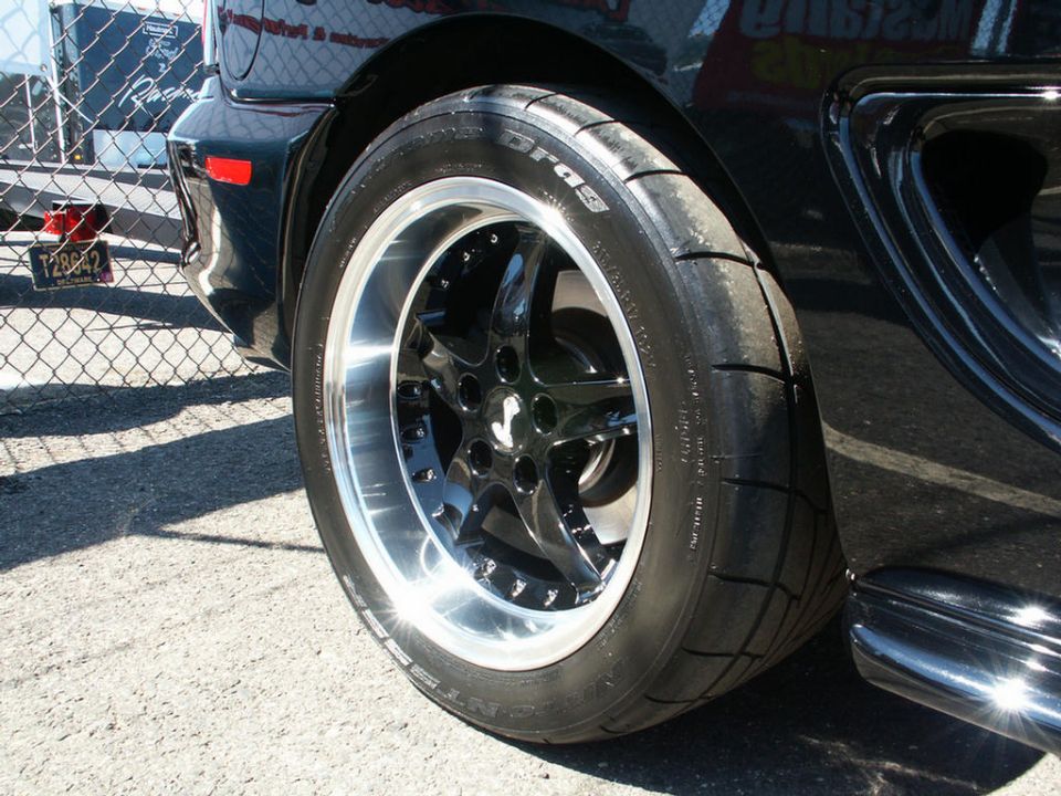 Close-up of a shiny black car wheel with a polished lip and a performance tire. A chrome trim piece is visible on the car's body