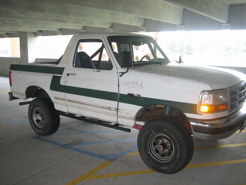 White Ford pickup truck with a green stripe down the side, parked in a garage. It has a roll cage inside