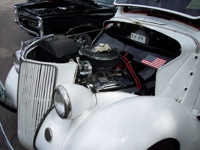 1972 Ford Ranchero - Engine bay of a white vintage car, an American flag sticker and a miniature car on the air filter, with another vehicle visible behind.