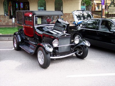 1972 Ford Ranchero - A black hot rod with its hood open is parked next to a black sedan on a street.