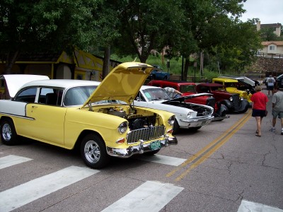 1972 Ford Ranchero - Several classic cars are lined up on a street, with one yellow and white Chevrolet prominently displayed with its hood open.