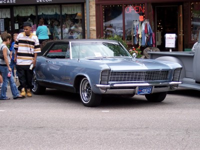 1972 Ford Ranchero - A blue 1967 Buick Riviera is parked on a street in front of shops, with a gray pickup truck to its right.