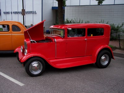 1972 Ford Ranchero - A bright red hot rod sedan sits with its hood up, parked next to an orange car.