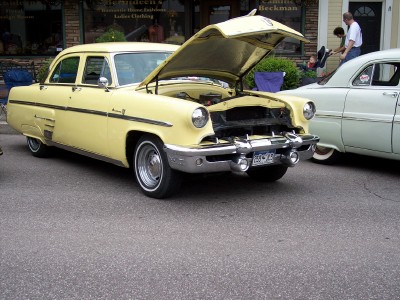 1972 Ford Ranchero - A yellow 1954 Ford Customline sedan is parked on a street with its hood open, next to another vintage car.