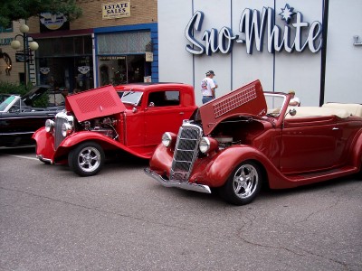 1972 Ford Ranchero - Two vintage red cars are displayed with their hoods open in front of a building with a "Snow-White" sign.