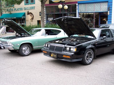1972 Ford Ranchero - Two cars with hoods open parked on a street in front of shops.