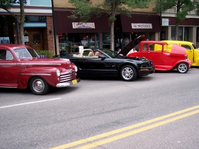 1972 Ford Ranchero - Several vehicles are parked in a line on a street in front of storefronts.