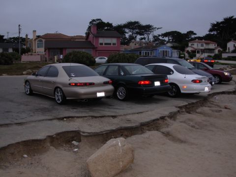 1995 Ford Taurus - A row of sedans and a minivan are parked near the coast, with houses in the background.