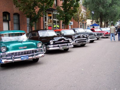 1972 Ford Ranchero - A row of classic cars, including a green 1955 Chevrolet, are parked along a street in front of brick buildings.