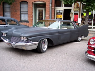 1972 Ford Ranchero - A matte black 1959 Chrysler Imperial with distinctive tail fins is parked on a street in front of brick buildings.