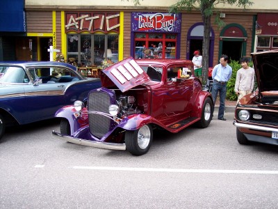 1972 Ford Ranchero - A custom coupe is displayed with its hood open, alongside other cars in front of shops.