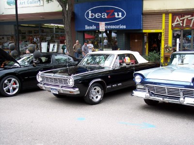 1972 Ford Ranchero - Three cars are parked in front of shops on a street, with people milling about.