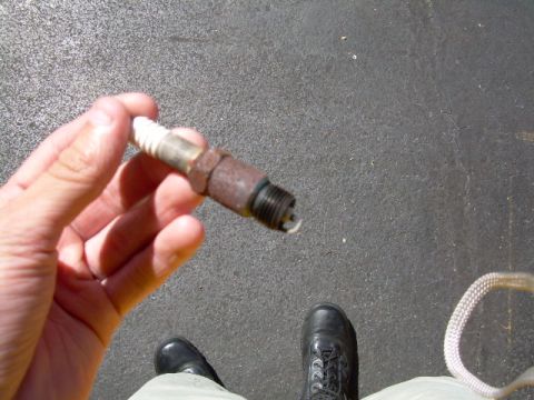 1971 Ford F-100 - A hand holds a rusty spark plug against a dark asphalt background, with feet in boots visible below.