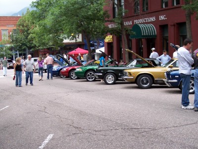 1972 Ford Ranchero - A row of classic cars, including a gold 1967 Ford Mustang, are displayed on a street with people milling around.