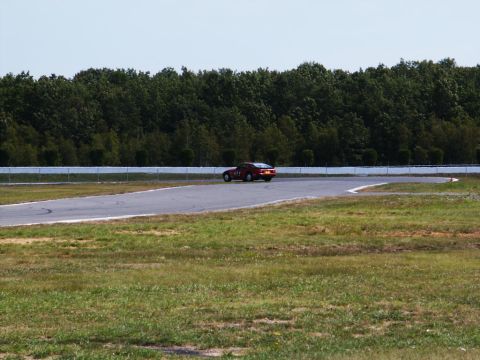 2001 Mercury Grand Marquis - A red race car navigates a turn on a paved track, with a dense line of trees bordering the course.