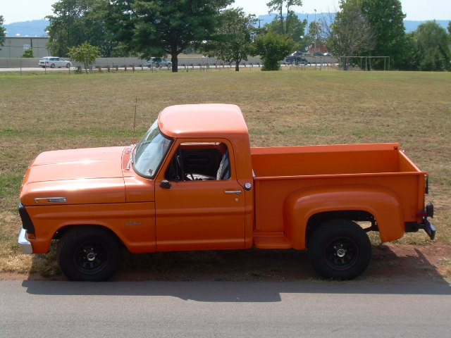 Side profile of an orange Ford stepside pickup with black wheels parked on pavement