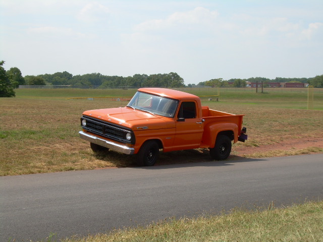 Front three-quarter of an orange Ford pickup with chrome bumper parked by a field