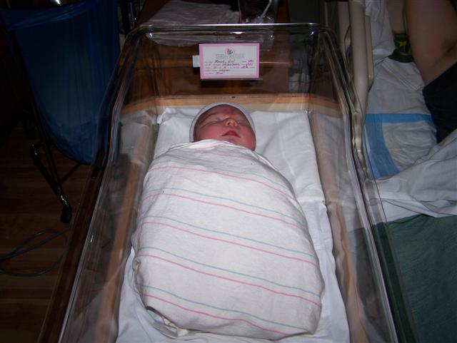 Newborn baby wrapped in a striped blanket, sleeping in a clear hospital bassinet. A pink card is attached to the side