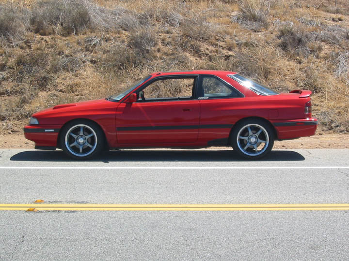 Red Mazda MX-6 coupe in profile on a desert highway, lowered on dark alloys