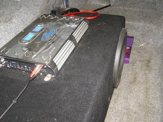 Car audio amplifier with blue lights and red/black RCA cables connected to a subwoofer with a purple component visible