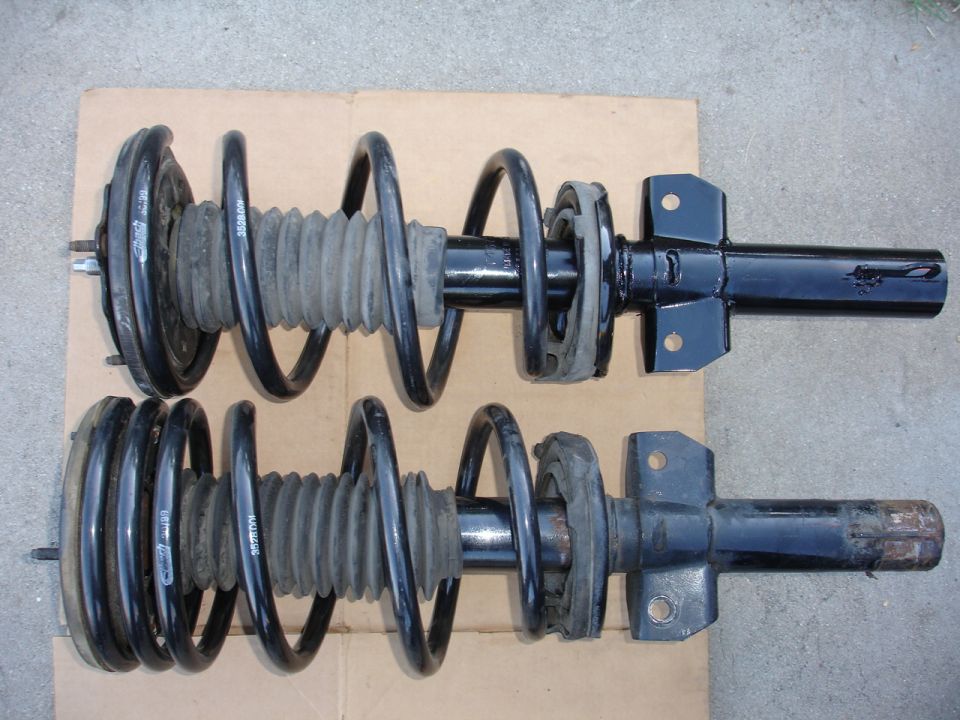 Two black front strut assemblies with coiled springs and rubber bellows are laid out on a cardboard surface