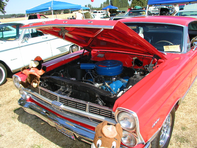 Red 1967 Ford Ranchero with hood open, showing blue engine components. Two stuffed animals are on the front