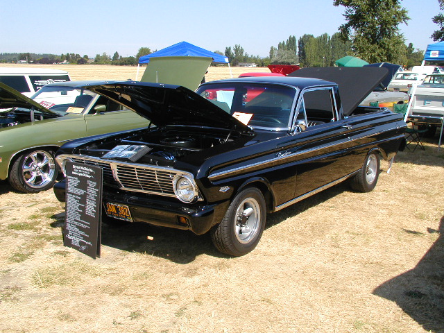 Black 1965 Ford Ranchero with hood open, parked on dry grass next to a green car