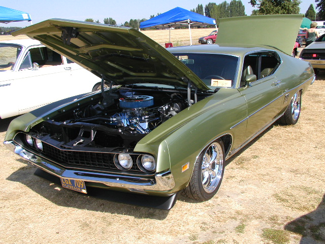 Green 1970 Ford Torino GT with hood open, showing engine. Another car is parked to its left