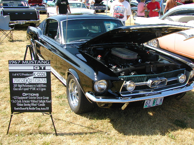 Black 1967 Ford Mustang GT with hood open, showing engine. Sign lists options and production numbers