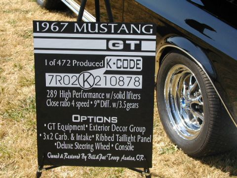 1967 Ford Ranchero - A sign displays details about a 1967 Mustang GT K-Code, including its VIN and options, next to a black car.