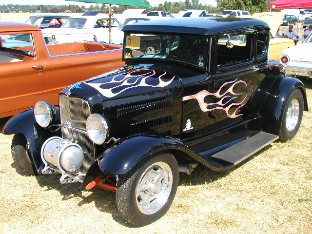 black hot rod coupe with flame decals on hood and side, parked on grass next to orange pickup truck