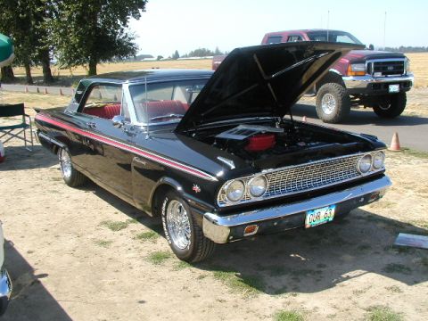 1967 Ford Ranchero - A black 1963 Ford coupe with a red interior is displayed with its hood open, parked outdoors next to a large truck.