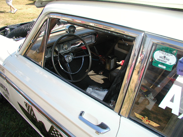 Interior of a white 1965 Ford Falcon with a black steering wheel and dashboard. A contest sticker is on the side window