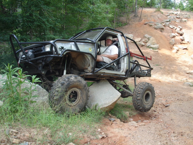 Stripped silver rock crawler with roll cage on big mud tires climbing a concrete block on a trail