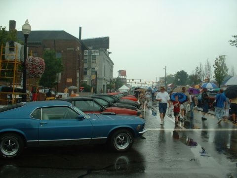 1978 Ford Mustang II - A blue coupe is parked on a wet street during an outdoor event with many people holding umbrellas.