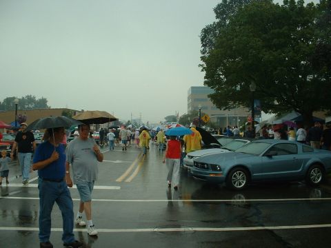 1978 Ford Mustang II - A blue coupe is parked on a wet street during a car show, with people holding umbrellas.
