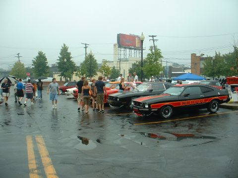 1978 Ford Mustang II - Row of Mustang IIs parked nose-in on a rain-soaked show lot, led at right by a black Cobra hatchback with orange "COBRA" side stripes and s…