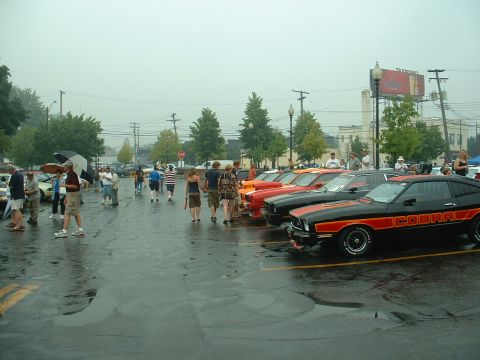 1978 Ford Mustang II - Several classic cars, including a black and orange Cobra, are parked in a lot on a rainy day.