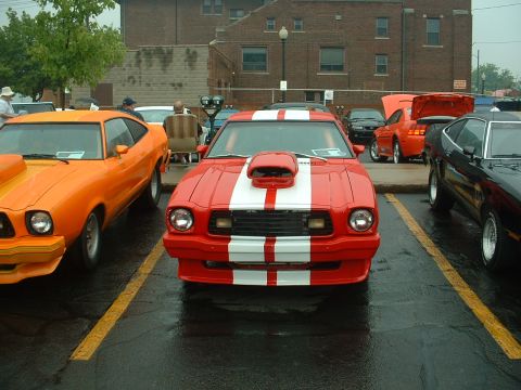1978 Ford Mustang II - Head-on front view of a red Mustang II Cobra II-style coupe with white dual racing stripes, front air dam and hood scoop, parked on wet asp…