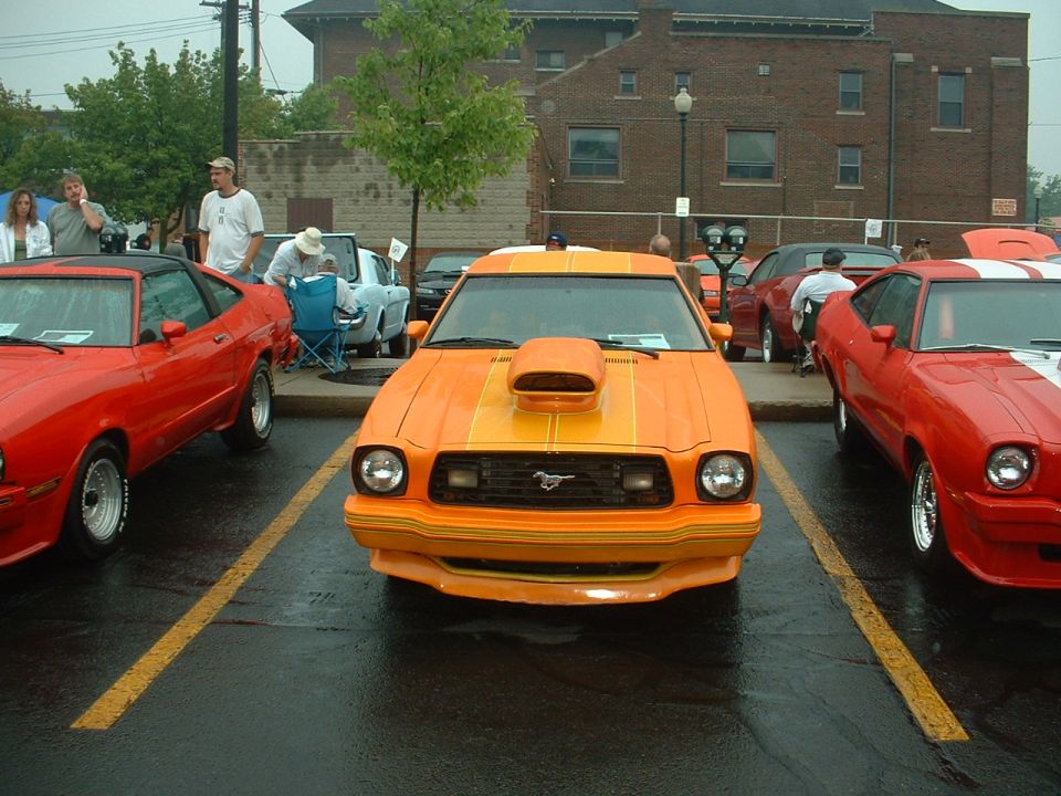 Orange Mustang II with hood scoop and stripes flanked by two red Mustang IIs at a show