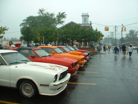 1978 Ford Mustang II - A line of classic cars, including a white coupe with gold graphics, are parked on a wet asphalt lot.