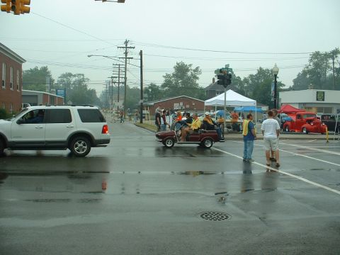 1978 Ford Mustang II - Rain-soaked street scene at a car show, with a white Ford Explorer crossing a wet intersection and a maroon golf cart carrying event staff…