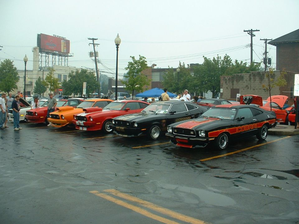 Black and orange 1975 Ford Mustang II with red trim and side stripes parked on a wet asphalt lot