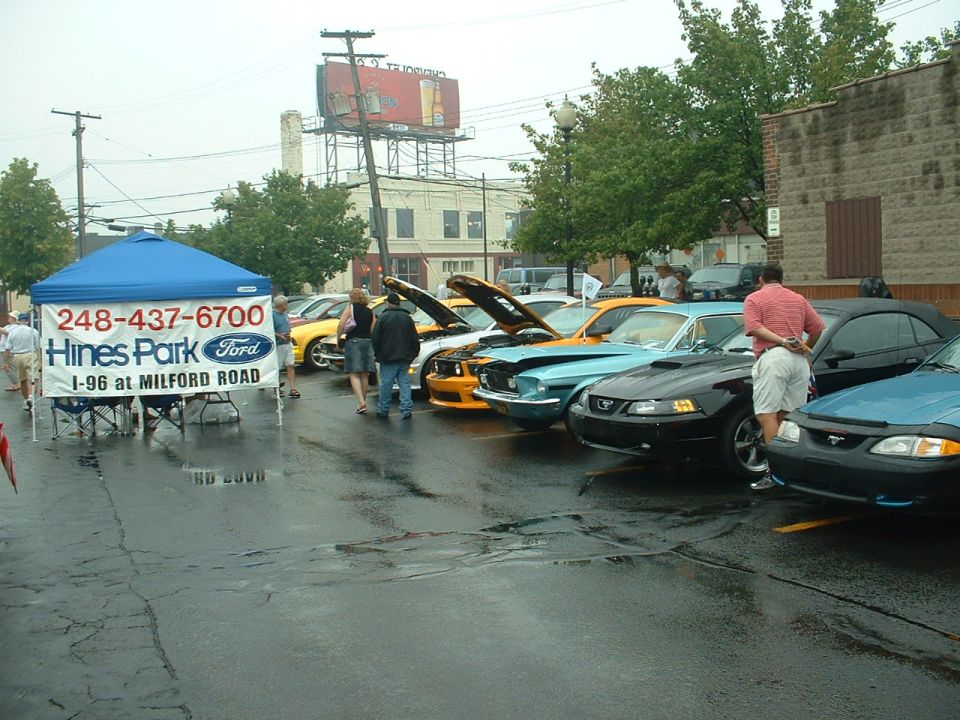 Black Ford Mustang with hood open, parked in a wet lot next to other Mustangs